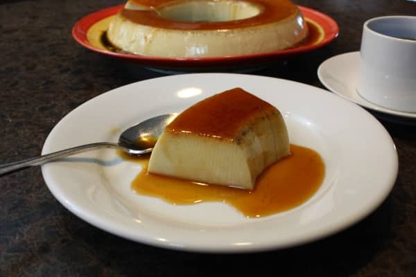 a piece of coconut flan in caramel sauce on a white plate with a spoon with the whole cake in the background