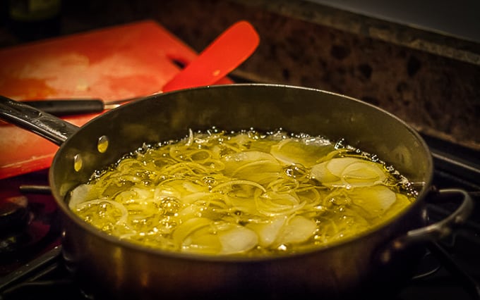 oil simmering in a pan with sliced potatoes and onions and a red cutting board in the background