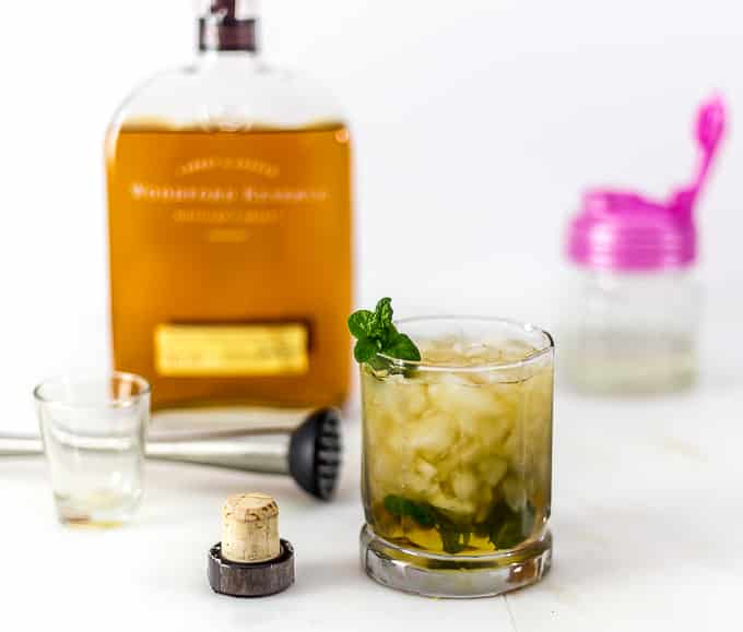 a mint julep in a glass next to a cap with bourbon, a muddler, and a shot glass in back