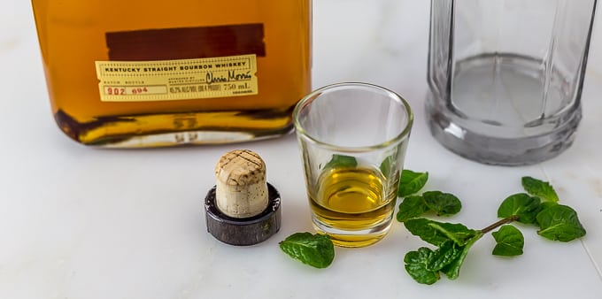 mint leaves, a shot glass with bourbon, and a cap with bourbon and a glass in back