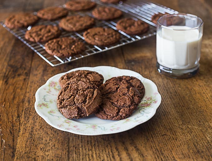 four chocolate cookies on an antique plate with a glass of milk on the right and a drying rack of cookies in the back