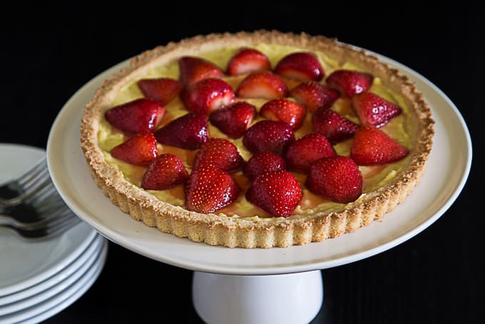 a large strawberry tart on a cake riser with a stack of plates and cutlery on the left