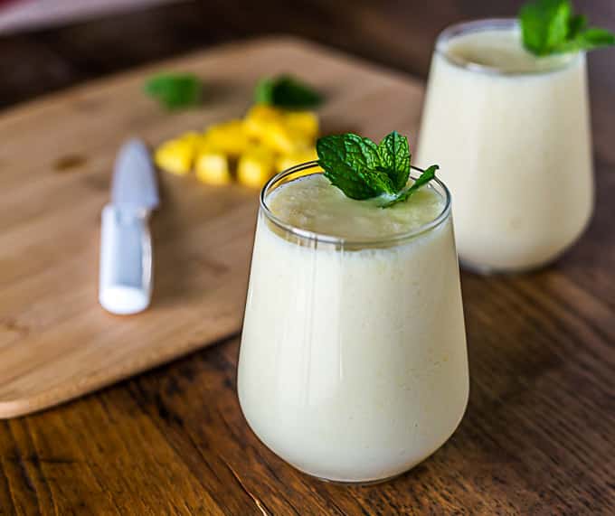 Mango lassi in glasses with mint leaves and a cutting board of mango in the back