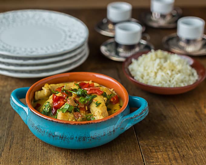 a blue pot of coconut chicken curry with a bowl of rice in the back and white plates on the back left
