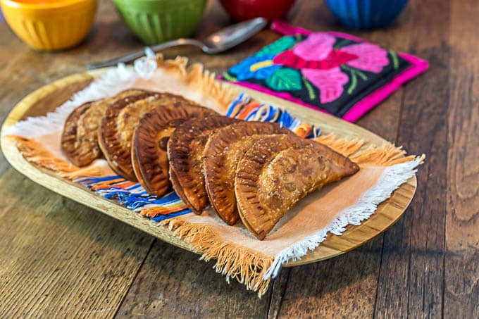 large fried empanadas on a wooden plate with colorful bowls in the back
