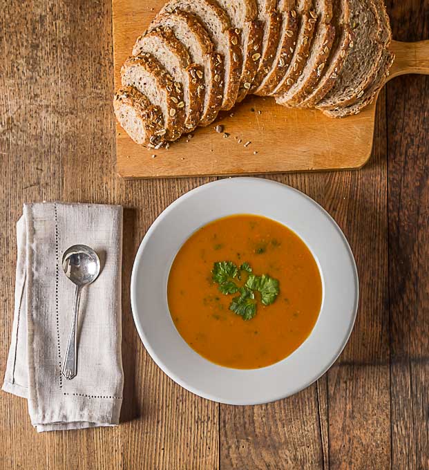 a bowl of carrot coriander soup with a spoon and napkin on the left and a loaf of multigrain bread above