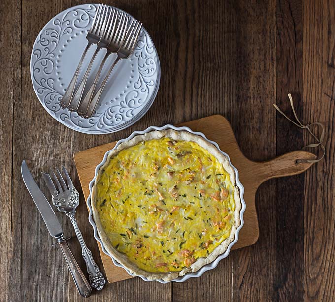 a salmon quiche on a cutting board with serving cutlery on the left and a stack of plates and forks above