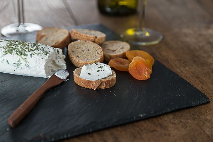 a log of herb goat cheese on a slate with a knife, sliced bread, and dried apricots