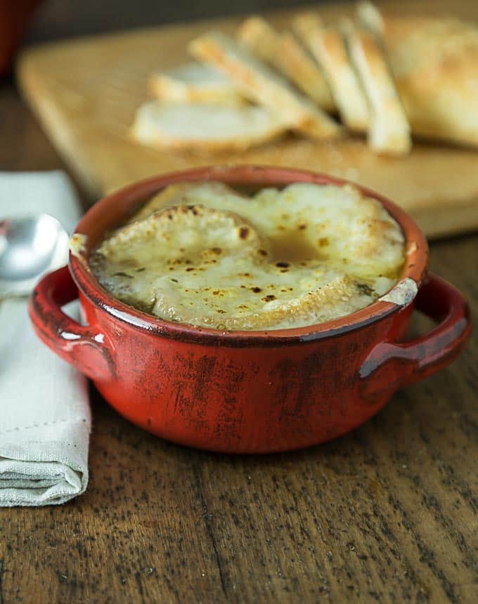 a close up red bowl of French onion soup with a sliced loaf of bread in the back