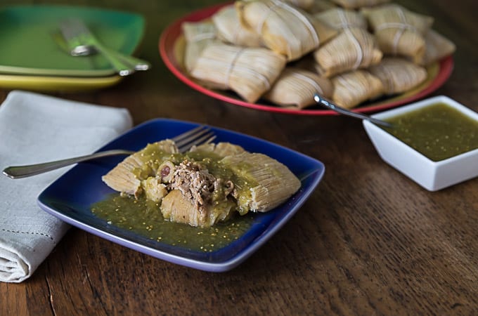 Pork tamales in salsa verde on a a blue plate with more tamales and sauce in the back