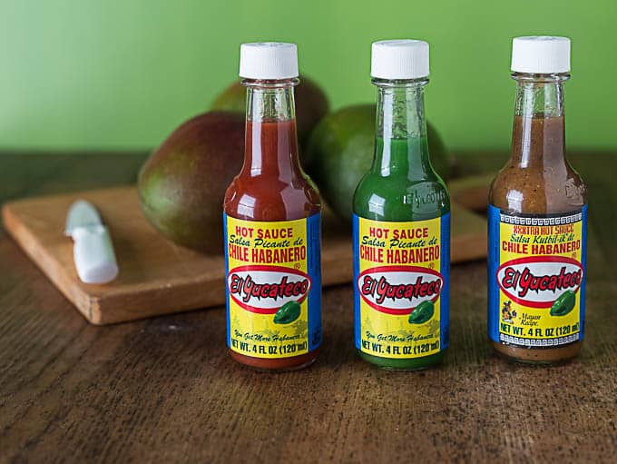 three bottles of el yucaterco hot sauce in a line with mangoes in the back