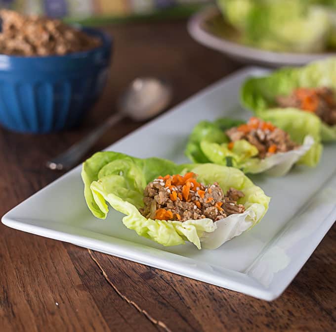 a white plate with three lettuce wraps on a wooden table and a bowl of meat and a spoon on the left.