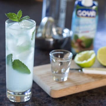 A photo of a coconut mojito in a clear tall glass with limes on a cutting board in the background.