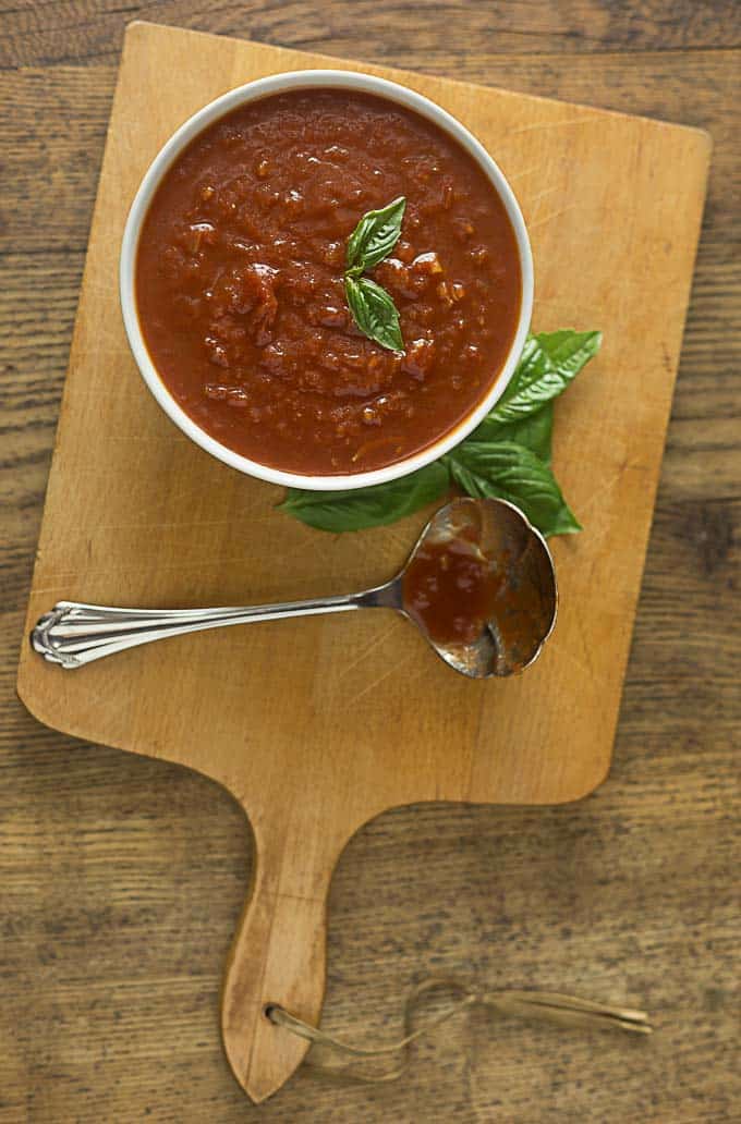 a closeup of a white bowl of marinara sauce with basil leaves and a silver ladle on a cutting board