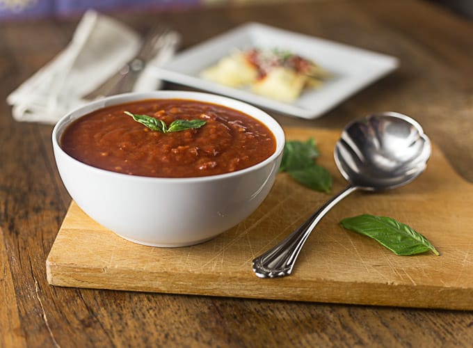a white bowl of marinara sauce next to a silver ladle on a cutting board