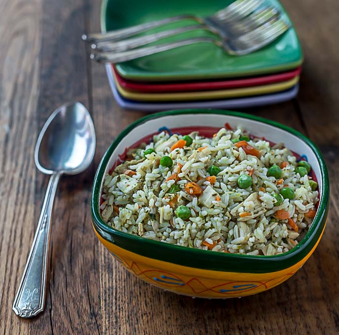 a bowl of rice with peas, turkey, and carrots on a wooden table with a spoon on the left