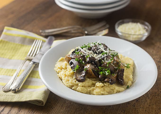 a white bowl of polenta and mushrooms with a fork, knife and yellow napkin on the left