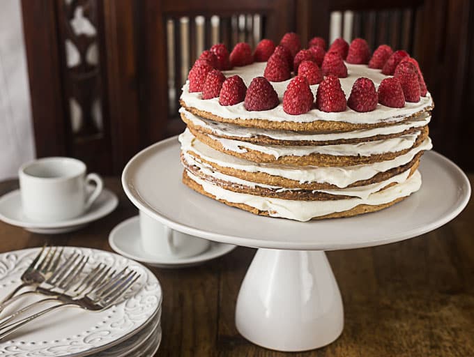 a swedish layered cake with raspberries on a bar next to a stack of white plates 