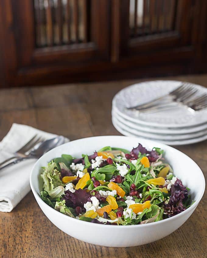 a white bowl of salad with purple lettuce, apricots, and feta cheese with a stack of plates in the back