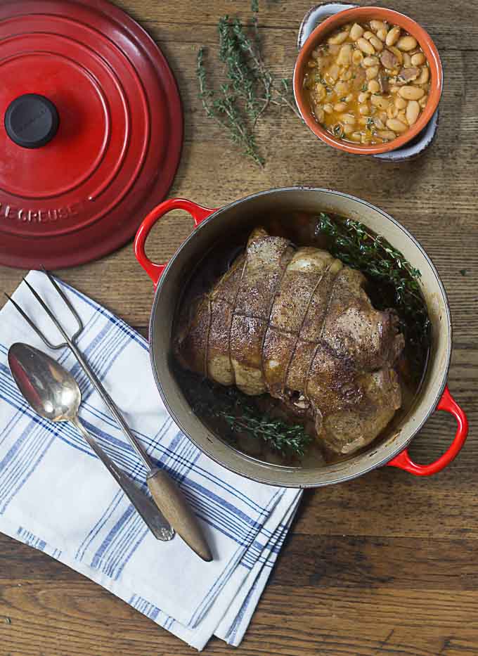 a herbed leg of lamb in a red dutch oven with a fork and spoon on a napkin on the left