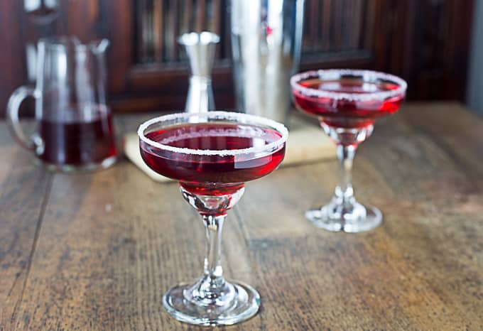 two pomegranate margaritas with salted rims on a wooden table with a bar in the back