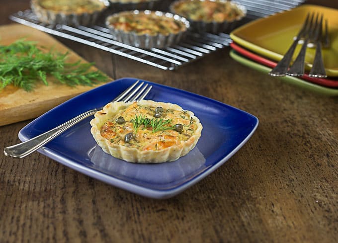 a smoked salmon tartlet on a blue plate with a fork and more tartlets on a drying rack in the back
