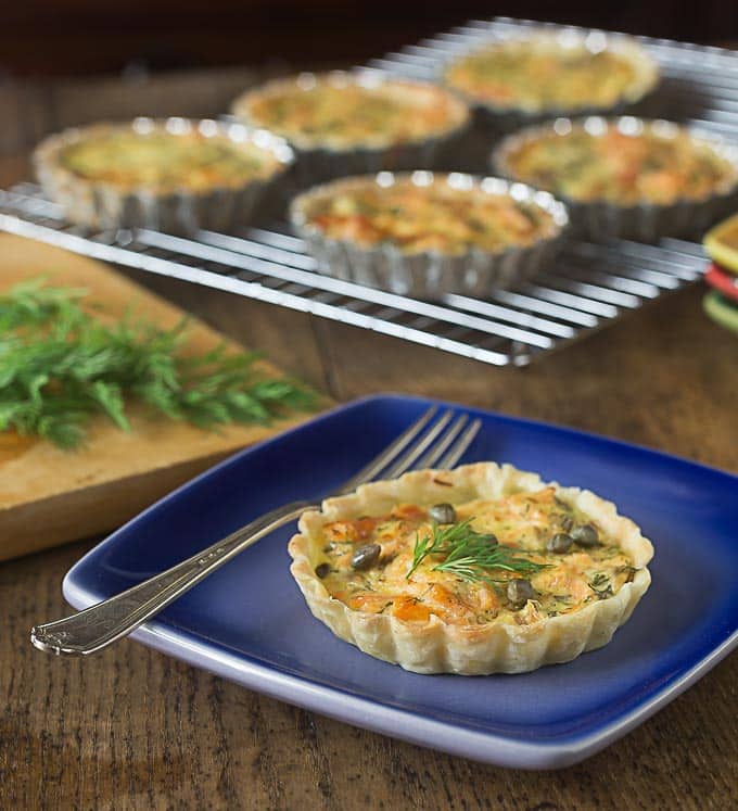 a closeup of a salmon tartlet on a plate with a fork and a drying rack in the back