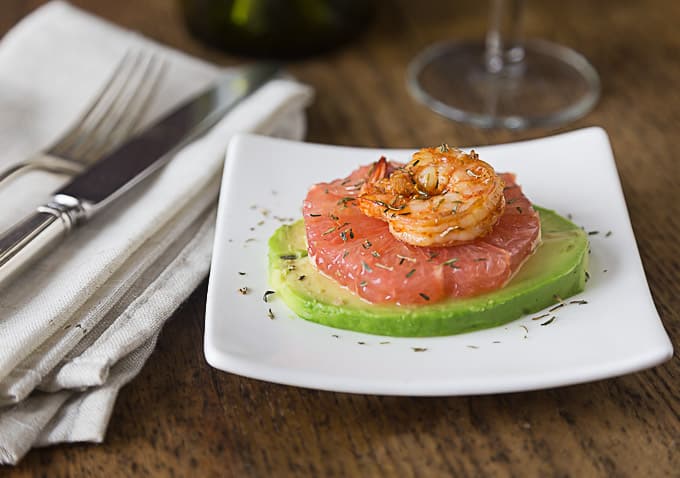 a white plate layered with avocado, grapefruit, and shrimp with a grey napkin and cutlery on the left