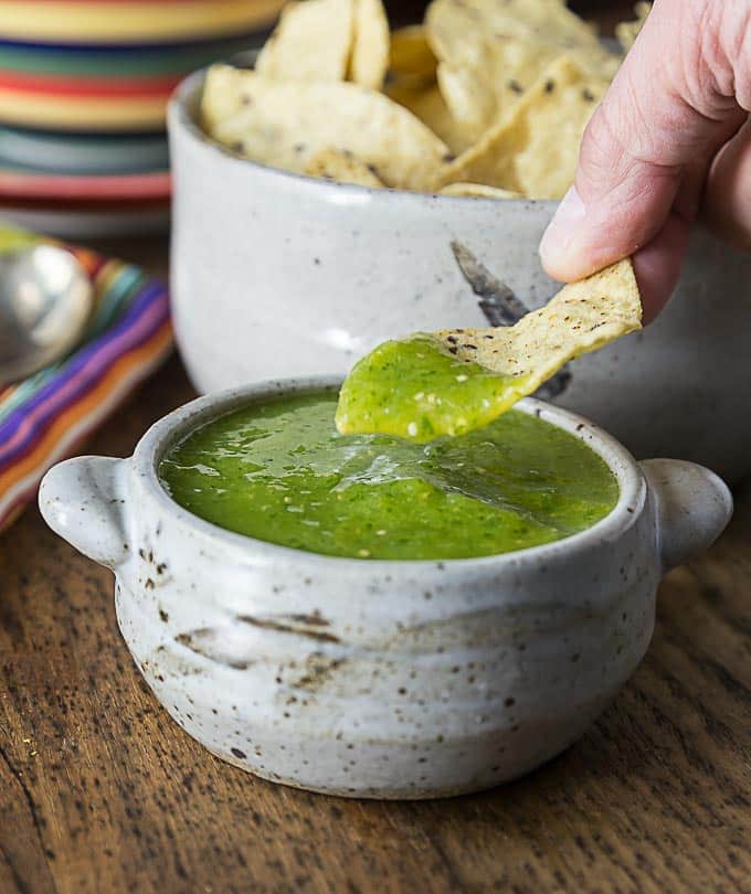 a wooden table with bowl of green salsa and a chip being dipped with a bowl of chips in the background