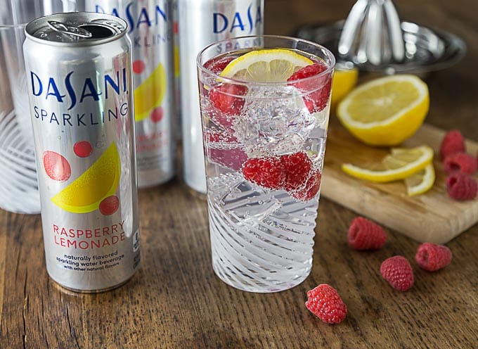 a glass of lemon berry mocktail next to cans of sparkling water with fruit on a cutting board