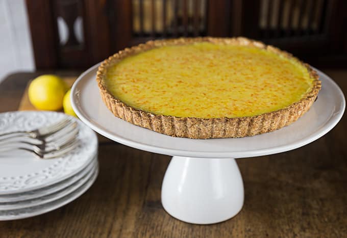 a lemon tart on a cake tray with a stack of plates and forks on the left