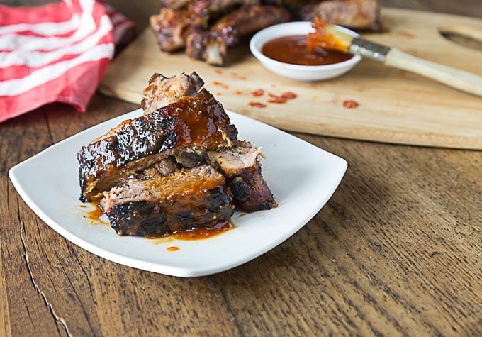 a plate of grilled ribs in bbq sauce with a cutting board of ribs and a brush in the background