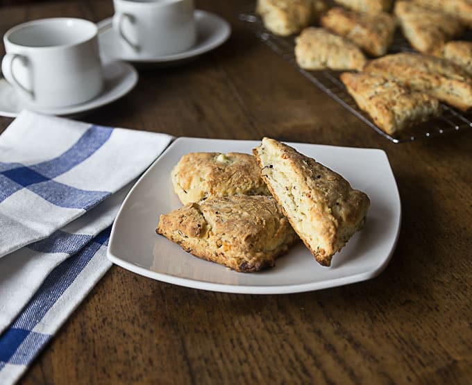 a white plate with three scones with a drying rack of scones and coffee cups in the back