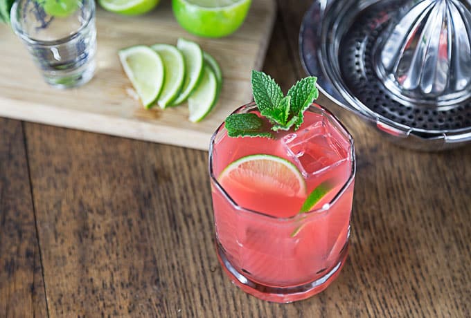 a pink cocktail in a glass with lime and mint with a juicer and sliced limes in back