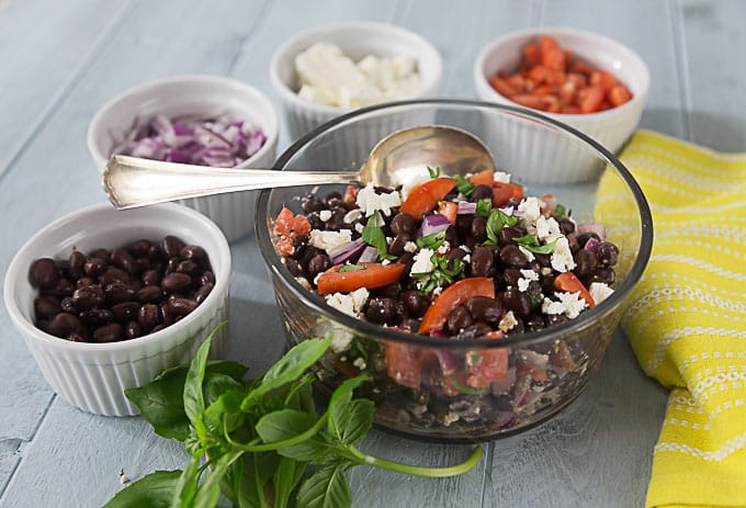 A bowl of black bean, feta and tomato salad with white bowls of ingredients all around the edge