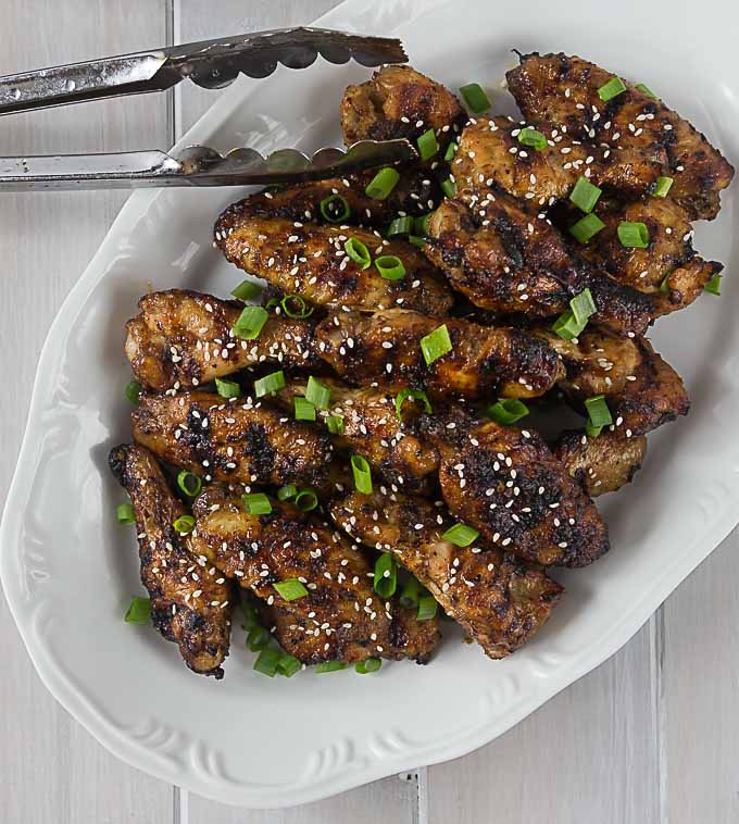 a platter of grilled chicken wings with scallion pieces and tongs