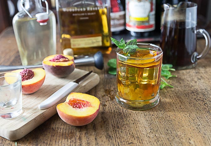 a wooden table with cut up peaches, a bourbon cocktail, and booze bottles