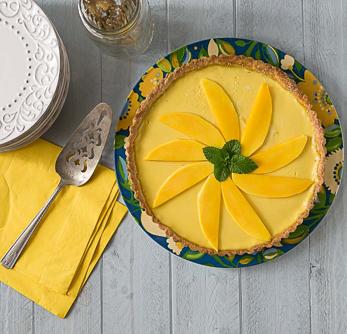 a mango passionfruit tart on a colored plate with a pie knife, yellow napkins, and white plates on the left