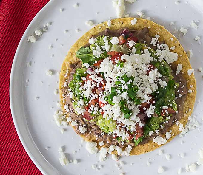 a tostada on a white plate with crumbled cheese, beans, lettuce, and tomato