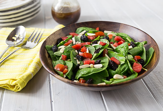 a spinach salad in a wooden bowl topped with tomatoes, raisins, and almonds with cutlery on the left