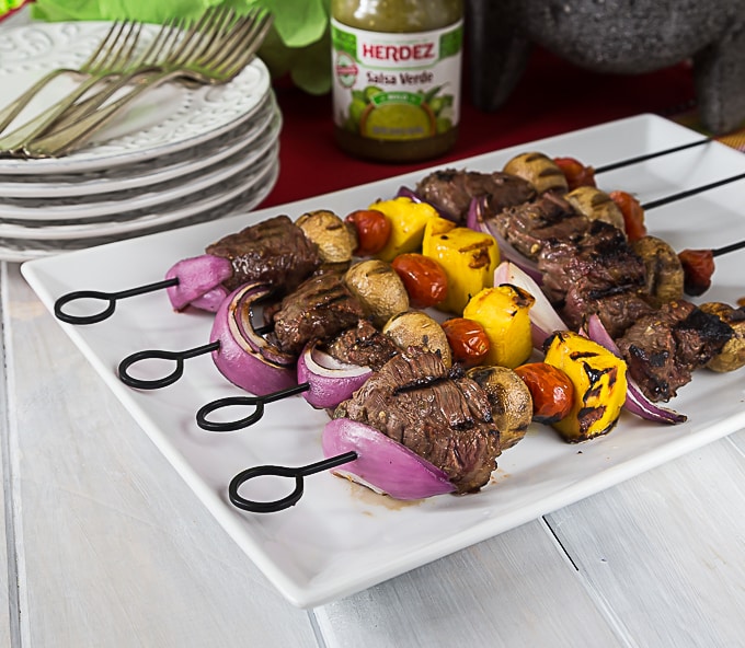 a white plate with beef kebabs on metal skewers with a stack of plates and forks in the back
