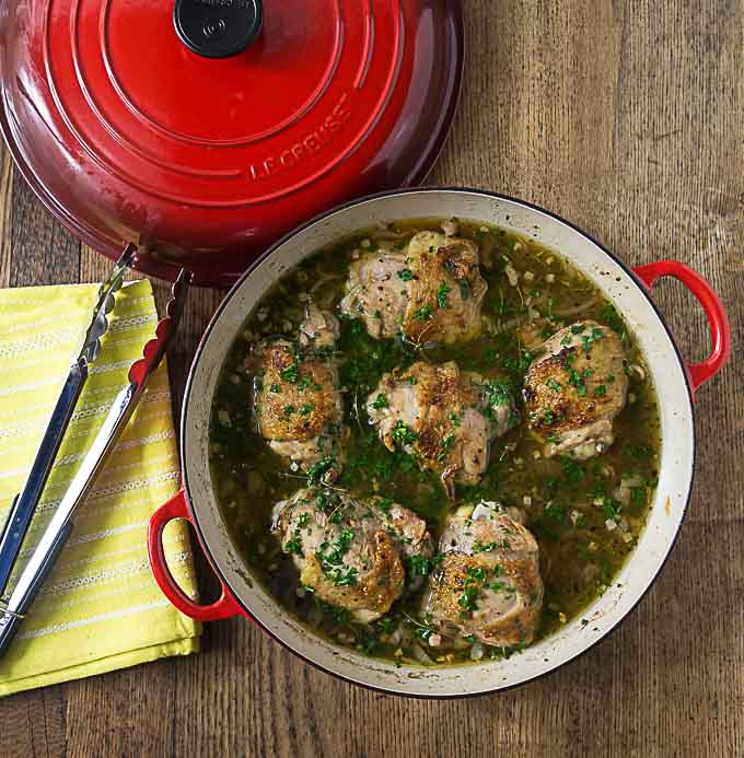 a red braising pan filled with chicken and juice topped with parsley with a set of tongs on the left