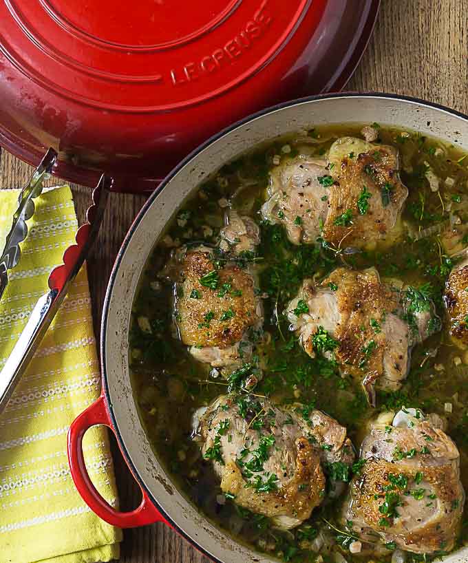 a red braising pan filled with chicken and juice topped with parsley 