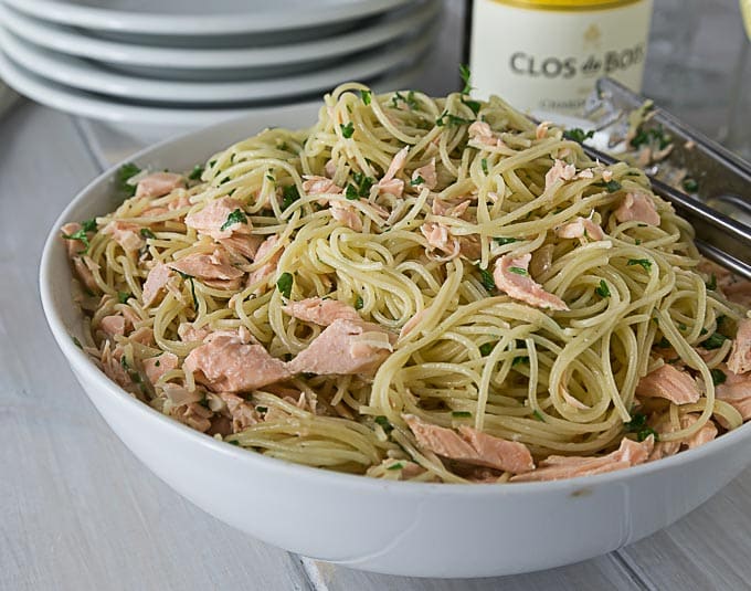 a white bowl with angel hair pasta and pieces of salmon with a bottle of wine in back