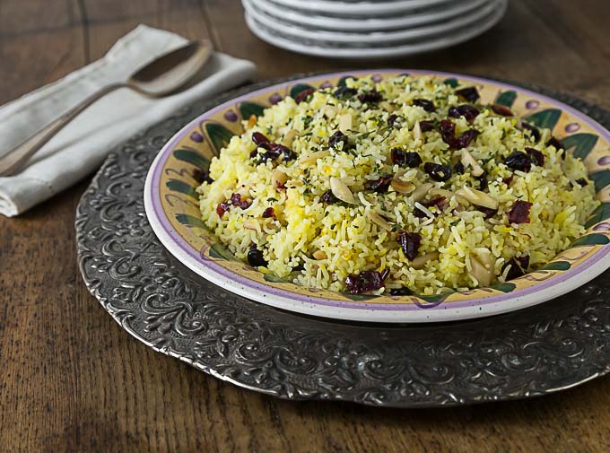 a sideview of a plate of saffron rice with craisins and almonds on a metal charger with a spoon on the left