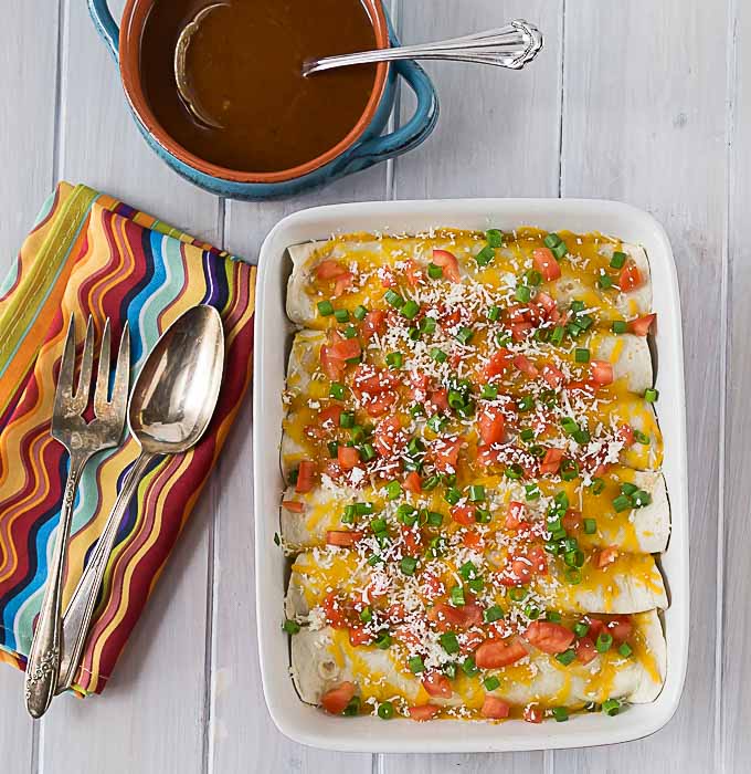 a tray of enchiladas with a colorful napkin on the left and a bowl of enchilada sauce on the top