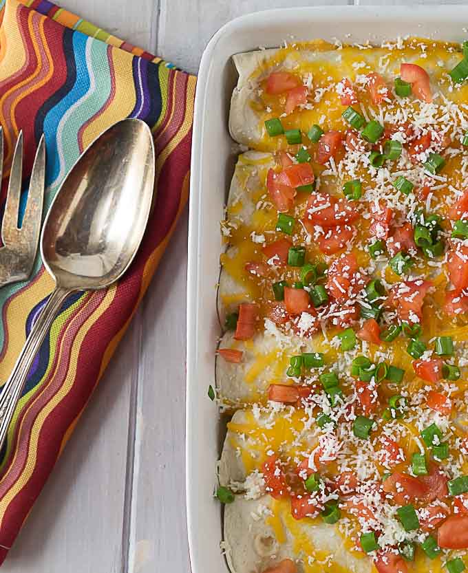 a close up of enchiladas with cheese, tomato, and green onion with a colorful napkin, spoon, and fork on the left