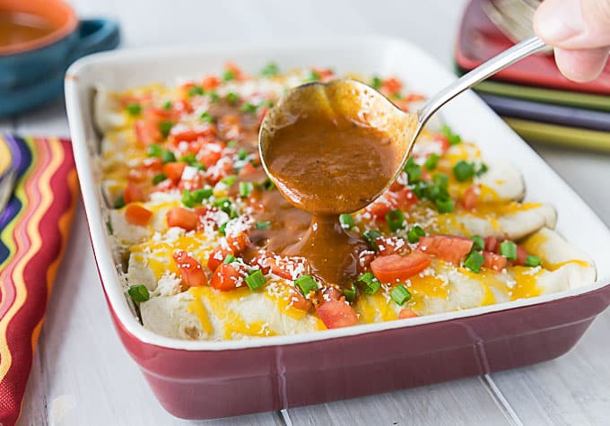 a tray of enchiladas topped with cheese, green onion, and tomato with a spoon pouring sauce over top
