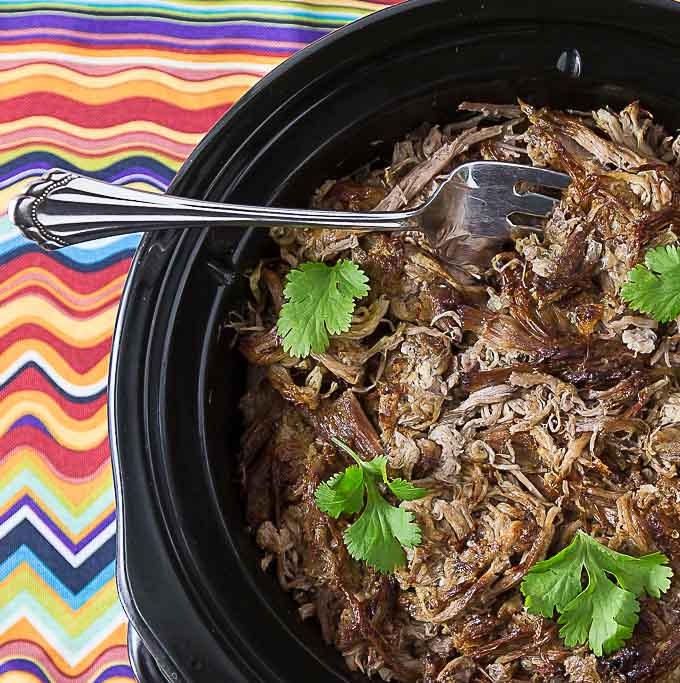 a crock filled with pork carnitas, coriander springs and a fork on a colorful napkin