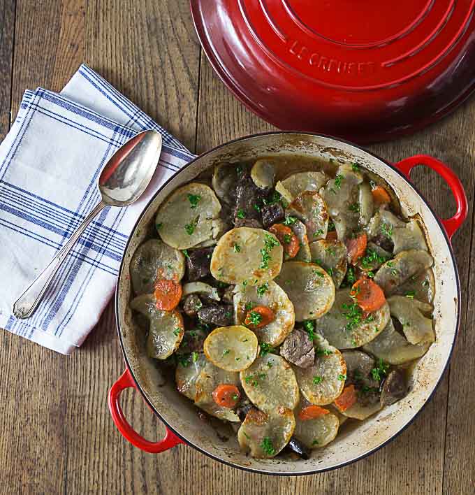 a dutch oven filled with potatoes, beef, and carrots with the lid on the table and a spoon and napkin on the left